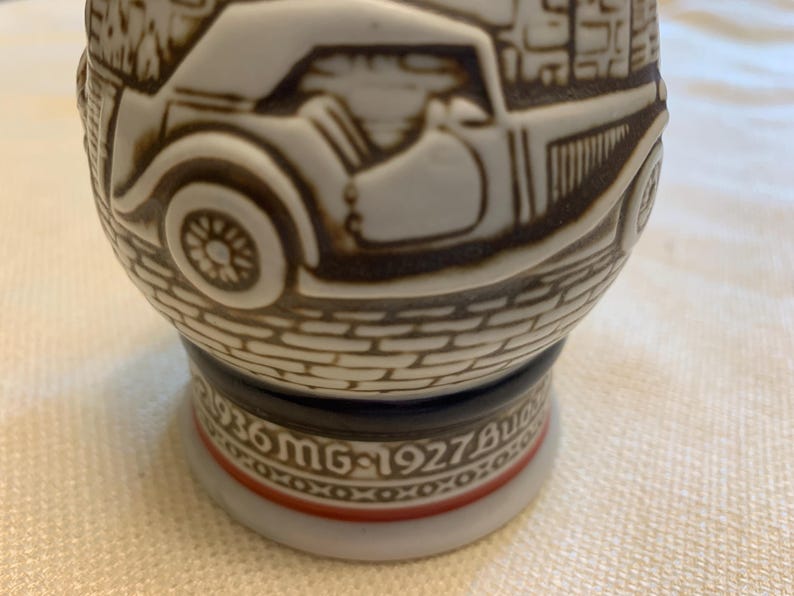 May include: A ceramic beer stein featuring a raised relief design of a vintage car on a cobblestone street. The base includes the text 1936 MG and 1927 Bugatti with a decorative red and black band. The stein has a white finish with brown detailing.