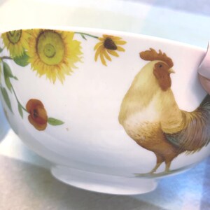 May include: A white ceramic bowl featuring a colorful illustration of a rooster standing among bright yellow sunflowers and small orange flowers. The design wraps around the exterior of the bowl, showcasing detailed feathers and green foliage.