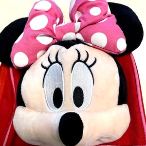 May include: A plush Minnie Mouse character head attached to a red and pink polka dot bag. The character features a large pink bow with white polka dots, black ears, embroidered eyes with long lashes, and a black nose on a light beige face.