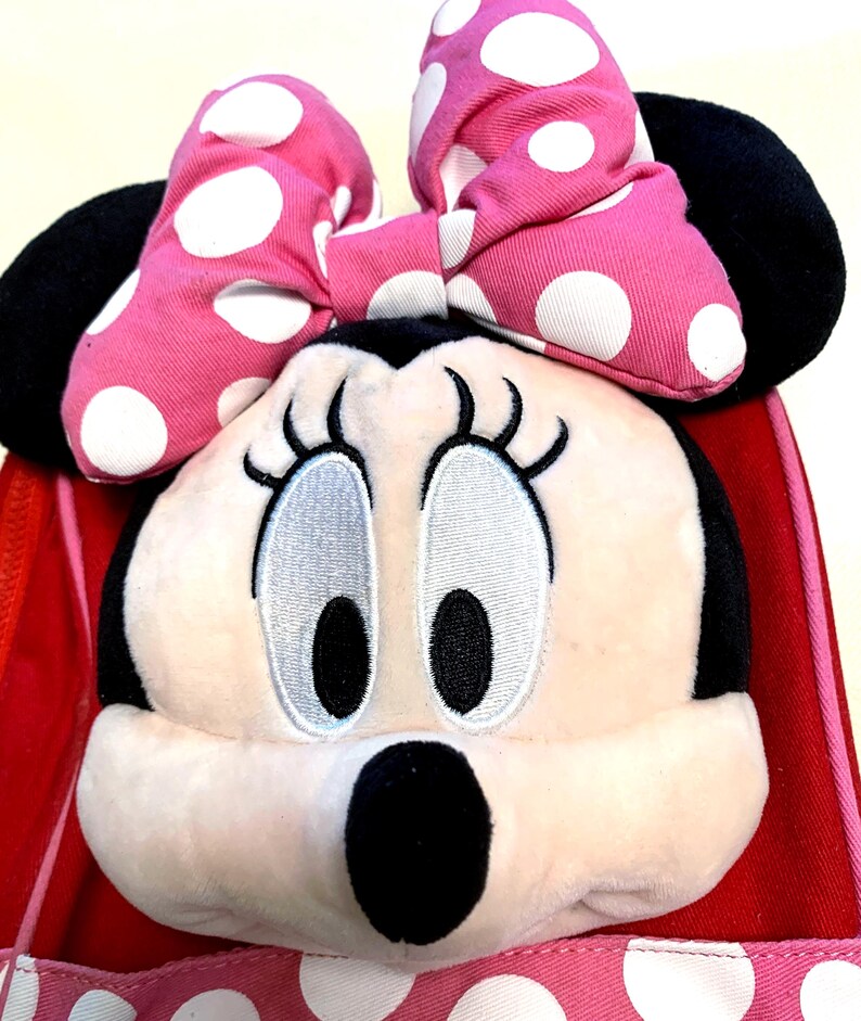 May include: A plush Minnie Mouse character head attached to a red and pink polka dot bag. The character features a large pink bow with white polka dots, black ears, embroidered eyes with long lashes, and a black nose on a light beige face.