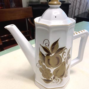 May include: A white ceramic teapot with an octagonal body and a matching lid featuring a brown finial. The exterior is decorated with a brown fruit motif including apples, pears, and grapes. It has a long spout and a handle with brown accents.