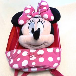 May include: A pink and red backpack featuring a plush Minnie Mouse head with a large pink and white polka dot bow. The front pocket is pink with white polka dots and displays the Disney logo embroidered in red thread on the front surface.