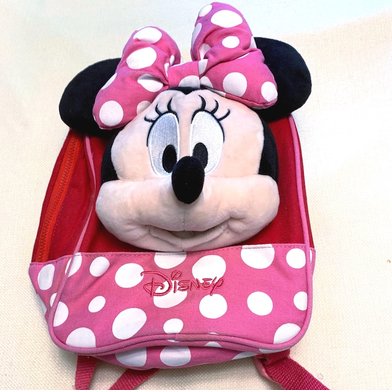 May include: A pink and red backpack featuring a plush Minnie Mouse head with a large pink and white polka dot bow. The front pocket is pink with white polka dots and displays the Disney logo embroidered in red thread on the front surface.