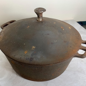 May include: A cast iron Dutch oven with a lid. The pot is dark brown with a slightly rusted appearance. It has two handles and a small knob on the lid. This cookware is suitable for cooking stews, soups, and other dishes.