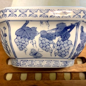 May include: A white ceramic bowl featuring blue hand-painted illustrations of grape clusters, leaves, and a butterfly. The rim has a geometric cross pattern, while the base is decorated with a Greek key border design in blue on a white background.