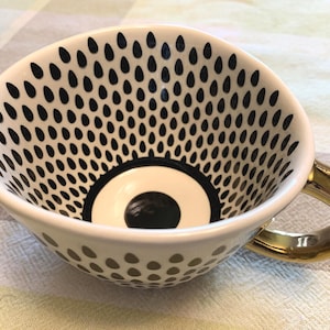 May include: A white ceramic teacup featuring a black teardrop pattern on the interior and exterior walls. The base of the cup interior has a solid black circle surrounded by a black ring. The cup includes a reflective gold-toned metallic handle.