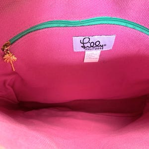 May include: Interior view of a pink tote bag with a green zipper and a gold palm tree charm. The bag features a label with the brand name "Lilly Pulitzer". The bag's interior is a vibrant pink color.
