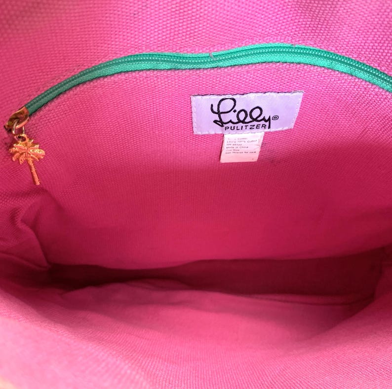 May include: Interior view of a pink tote bag with a green zipper and a gold palm tree charm. The bag features a label with the brand name "Lilly Pulitzer". The bag's interior is a vibrant pink color.