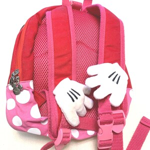 May include: A red and pink polka dot backpack featuring two plush white cartoon-style gloves attached to the back panel. The bag includes adjustable pink shoulder straps with mesh padding, a top handle, and a silver zipper pull shaped like a mouse head.