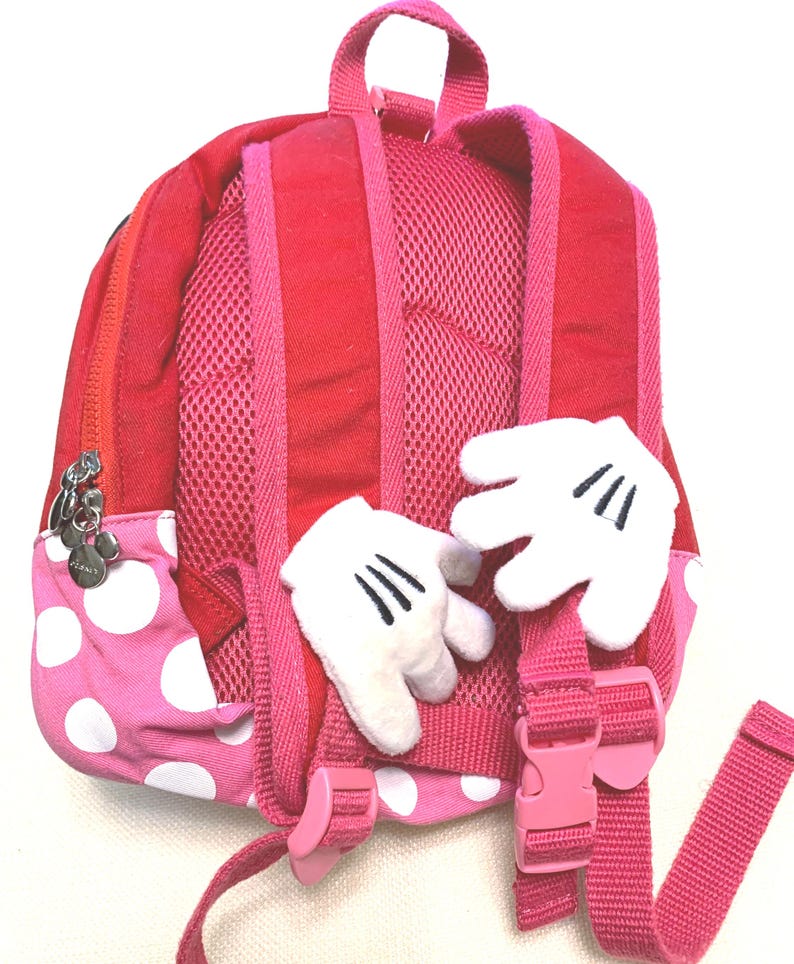 May include: A red and pink polka dot backpack featuring two plush white cartoon-style gloves attached to the back panel. The bag includes adjustable pink shoulder straps with mesh padding, a top handle, and a silver zipper pull shaped like a mouse head.