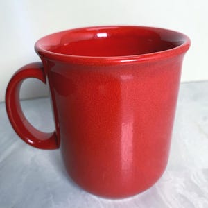 May include: A red ceramic mug with a rounded body and a curved handle. The mug has a glossy finish and a slightly flared rim. The mug is designed for hot beverages.