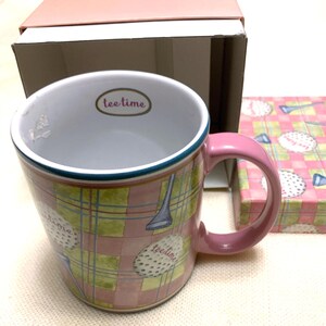 May include: A ceramic mug with a pink handle and a plaid pattern in shades of pink and green. The exterior features golf ball and club illustrations with the text "tee time" printed on the balls and inside the rim near the top edge.
