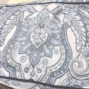 May include: A rectangular pillow sham featuring a woven fabric with a light blue, white, and dark blue paisley pattern. The edges are finished with a dark blue twisted rope cord trim that frames the intricate floral and leaf motifs throughout.
