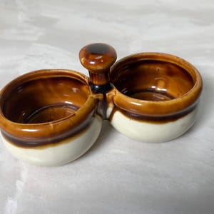 May include: A ceramic salt and pepper cellar with two connected bowls and a handle. The bowls are brown on the inside and have a white base with a brown stripe. The handle is brown.
