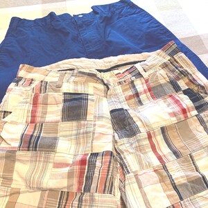 May include: A pair of solid blue shorts positioned behind a pair of shorts featuring a multi-colored plaid patchwork pattern. Both pairs of shorts include a waistband with a button closure, belt loops, and side pockets, showcasing a classic design.