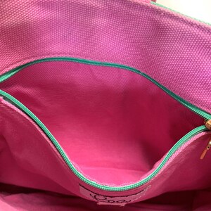 May include: Close-up of a pink tote bag interior with a zippered pocket. The bag has a green trim and a pink lining. The zipper is gold-colored. The bag has a label with cursive text.
