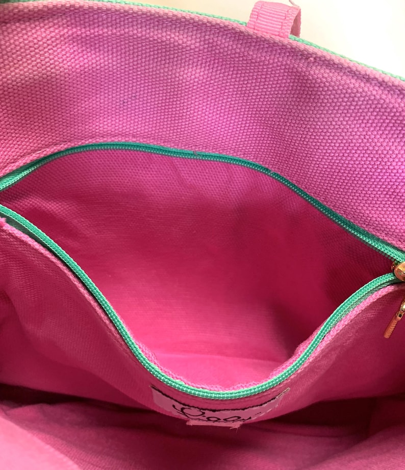May include: Close-up of a pink tote bag interior with a zippered pocket. The bag has a green trim and a pink lining. The zipper is gold-colored. The bag has a label with cursive text.
