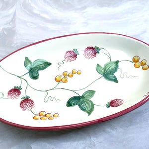 May include: Oval ceramic serving platter with a cream-colored surface, adorned with hand-painted red berries, green leaves, and yellow berries. The rim is a deep red. A decorative piece for serving food.