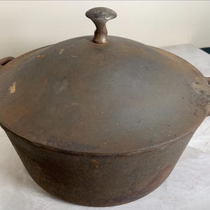 May include: A dark brown, weathered cast iron Dutch oven with a matching lid. The pot features two side handles with open rectangular cutouts and a central knob on the lid. The surface shows signs of oxidation and wear with visible rust spots.