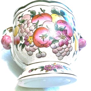May include: A ceramic bowl with a crackle glaze finish, featuring a colorful painted motif of various fruits including grapes, apples, and peaches. The bowl has two side handles shaped like small pink fruits and a floral pattern on the base.