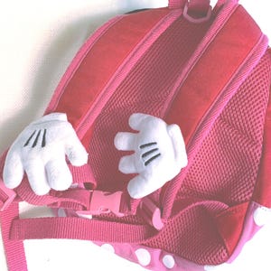 May include: A pink backpack featuring two white plush cartoon-style hands attached to the back panel. The backpack includes pink adjustable shoulder straps with a plastic buckle closure and a mesh-covered back padding for comfort and support.