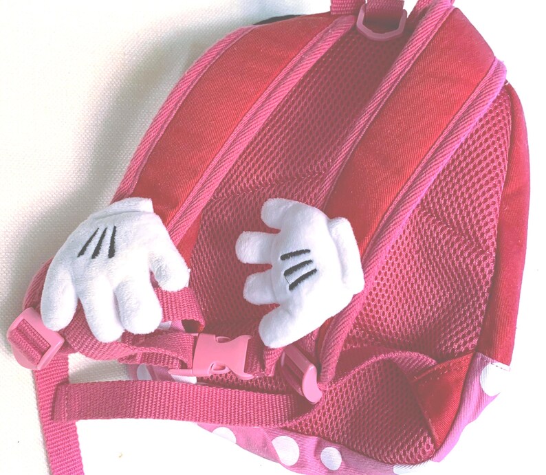 May include: A pink backpack featuring two white plush cartoon-style hands attached to the back panel. The backpack includes pink adjustable shoulder straps with a plastic buckle closure and a mesh-covered back padding for comfort and support.