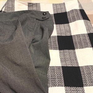 May include: A pair of solid charcoal gray trousers with two dark buttons at the waistband, positioned next to a skirt featuring a large black and white checkered pattern with a textured, woven fabric appearance and a straight hemline.