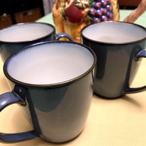 May include: Three blue ceramic mugs with rounded handles. The mugs have a gradient effect, transitioning from a light blue interior to a darker blue exterior. The rims are outlined with a thin brown line.
