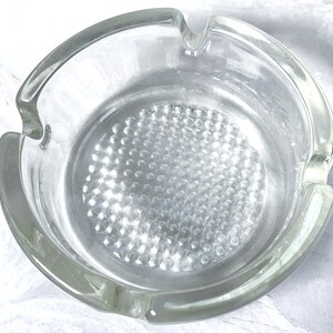 May include: A clear glass ashtray with a circular design. The ashtray has a rounded shape with a series of small indentations in the center. It features three notches for resting cigarettes. The glass has a slight green tint.