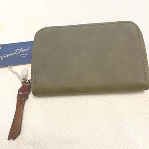 May include: An olive green rectangular wallet with a zipper closure and a brown leather pull tab. A blue rectangular tag attached to the zipper features the white text Universal Thread Goods Co. The wallet has a smooth, matte finish.