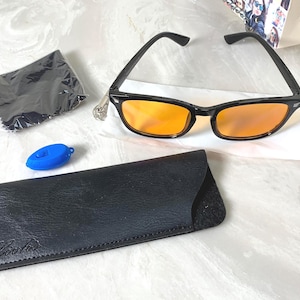 May include: Black-framed sunglasses with orange-tinted lenses, a black case, a blue accessory, and a black cleaning cloth. The sunglasses are on a white surface. The case has a logo on it.