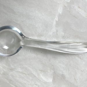 May include: A clear glass spoon with a rounded bowl and a long, curved handle. The spoon is made of transparent glass, allowing light to pass through. The design is simple and elegant, suitable for serving or decorative purposes.