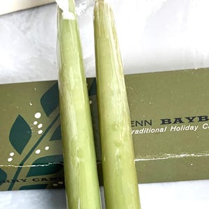 May include: Two green taper candles in plastic wrap, next to a green box with white text. The box has a leaf design and the text "ENN BAYB" and "traditional Holiday Ca".