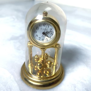May include: A gold-colored quartz clock with a white face and black numerals is encased in a clear glass dome. The clock has a round face with the word "CIRCA" above the hands. The clock sits on a gold-colored base with decorative elements.