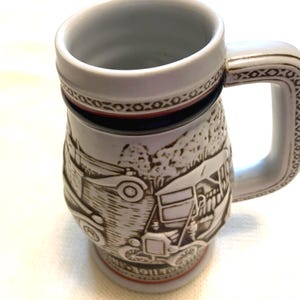 May include: A ceramic beer stein featuring a raised relief design of vintage automobiles. The mug has a white base color with brown detailing, a sturdy handle with a geometric pattern, and a red band near the base and rim. The surface is textured.
