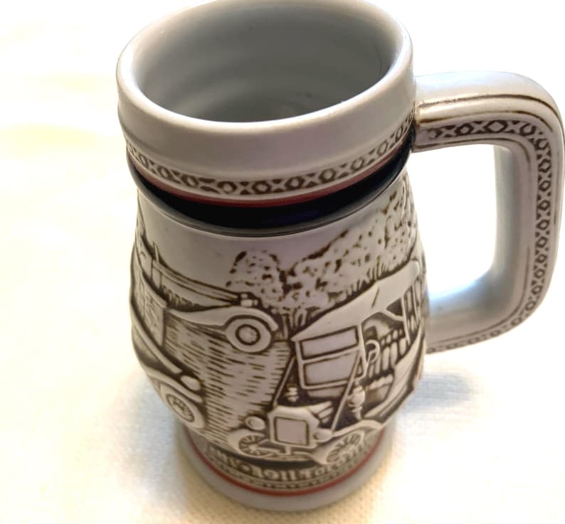 May include: A ceramic beer stein featuring a raised relief design of vintage automobiles. The mug has a white base color with brown detailing, a sturdy handle with a geometric pattern, and a red band near the base and rim. The surface is textured.