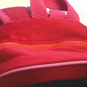 May include: A bright red fabric backpack featuring a prominent orange zipper closure that runs horizontally across the center. The bag includes a pink fabric trim along the bottom edge and a matching pink woven handle attached to the top section.
