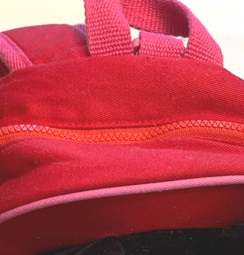 May include: A bright red fabric backpack featuring a prominent orange zipper closure that runs horizontally across the center. The bag includes a pink fabric trim along the bottom edge and a matching pink woven handle attached to the top section.