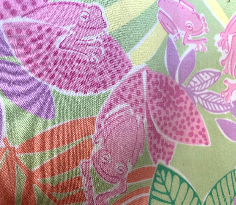 May include: A close-up of a fabric with a repeating pattern of pink frogs and leaves. The frogs are a solid pink color with darker pink spots. The leaves are green, orange, and purple. The background is a light green and yellow.