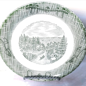 May include: A round, white and green decorative plate. The plate features a detailed black and white rural landscape scene in the center, surrounded by a green textured border and decorative elements.