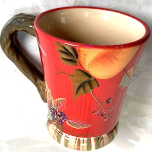 May include: A red ceramic mug with a tree branch handle. The mug features a fruit and leaf design in shades of orange, green, and purple. The base has a striped pattern. The mug is approximately 5 inches tall.
