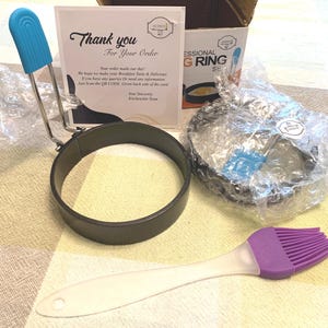 May include: A set of black metal egg rings with blue silicone handles, a purple silicone basting brush, a cardboard box labeled Professional Egg Ring Set, and a white thank you card with black text, all partially wrapped in clear bubble wrap.