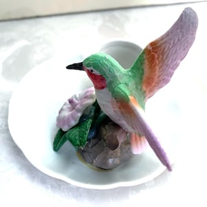 May include: A ceramic hummingbird figurine with outstretched wings, perched on a small rock with a flower. The bird features green, red, orange, and purple coloring. The figurine sits on a white, scalloped-edge saucer.