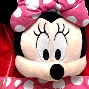 May include: A plush Minnie Mouse character head with black ears and a large pink bow featuring white polka dots. The face is cream-colored with embroidered eyes and eyelashes. Below the head is a pink fabric section with white polka dots and the red Disney logo.