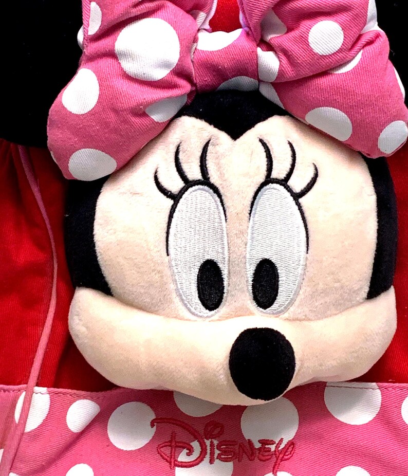 May include: A plush Minnie Mouse character head with black ears and a large pink bow featuring white polka dots. The face is cream-colored with embroidered eyes and eyelashes. Below the head is a pink fabric section with white polka dots and the red Disney logo.