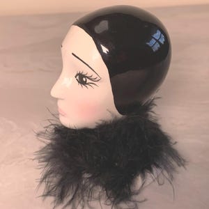 May include: A ceramic figurine of a woman's head, featuring a black, glossy bob hairstyle and a white face with painted features. The figurine has a black feather boa around the neck.