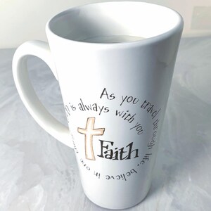 May include: White ceramic coffee mug with a curved handle. The mug features a cross and the word "Faith" in a handwritten font, surrounded by the phrase "As you travel through life, believe in one who is always with you."