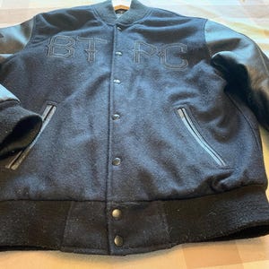 May include: A black varsity jacket featuring a wool-blend body and black leather sleeves. The front displays the embroidered letters BT PC in a gothic font, a snap-button closure, two side pockets with leather trim, and ribbed knit cuffs and hem.