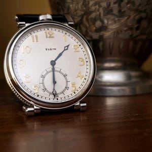 May include: A vintage Elgin pocket watch with a silver case and a white face. The watch features gold-coloured numerals and hands, with a smaller seconds dial. The watch is set on a dark wooden surface.