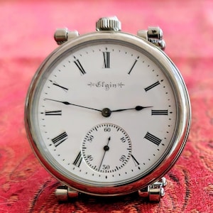 May include: A silver-toned Elgin pocket watch with a white dial, black Roman numerals, and thin black hands. The watch features a small sub-dial for seconds at the bottom and a winding crown at the top, with decorative metal lugs on the sides.